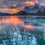 Reflective ice on a pond, mountains in the background, with a purple, orange, yellow sunset