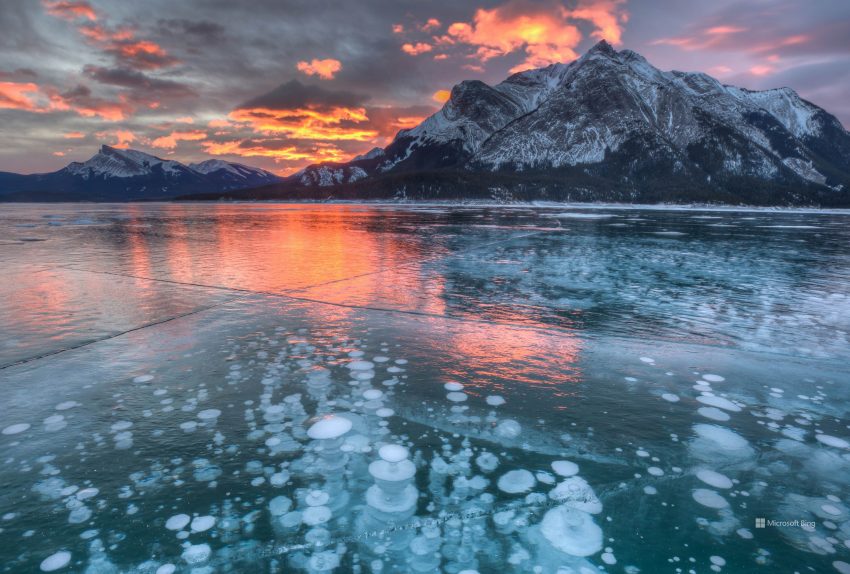 Reflective ice on a pond, mountains in the background, with a purple, orange, yellow sunset